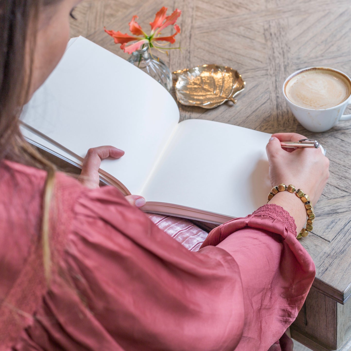 on a coffee table is a wedding guest book which is open, you can see the blank pages and woman is set to write a message in it. There is also a cup of coffee on the table 