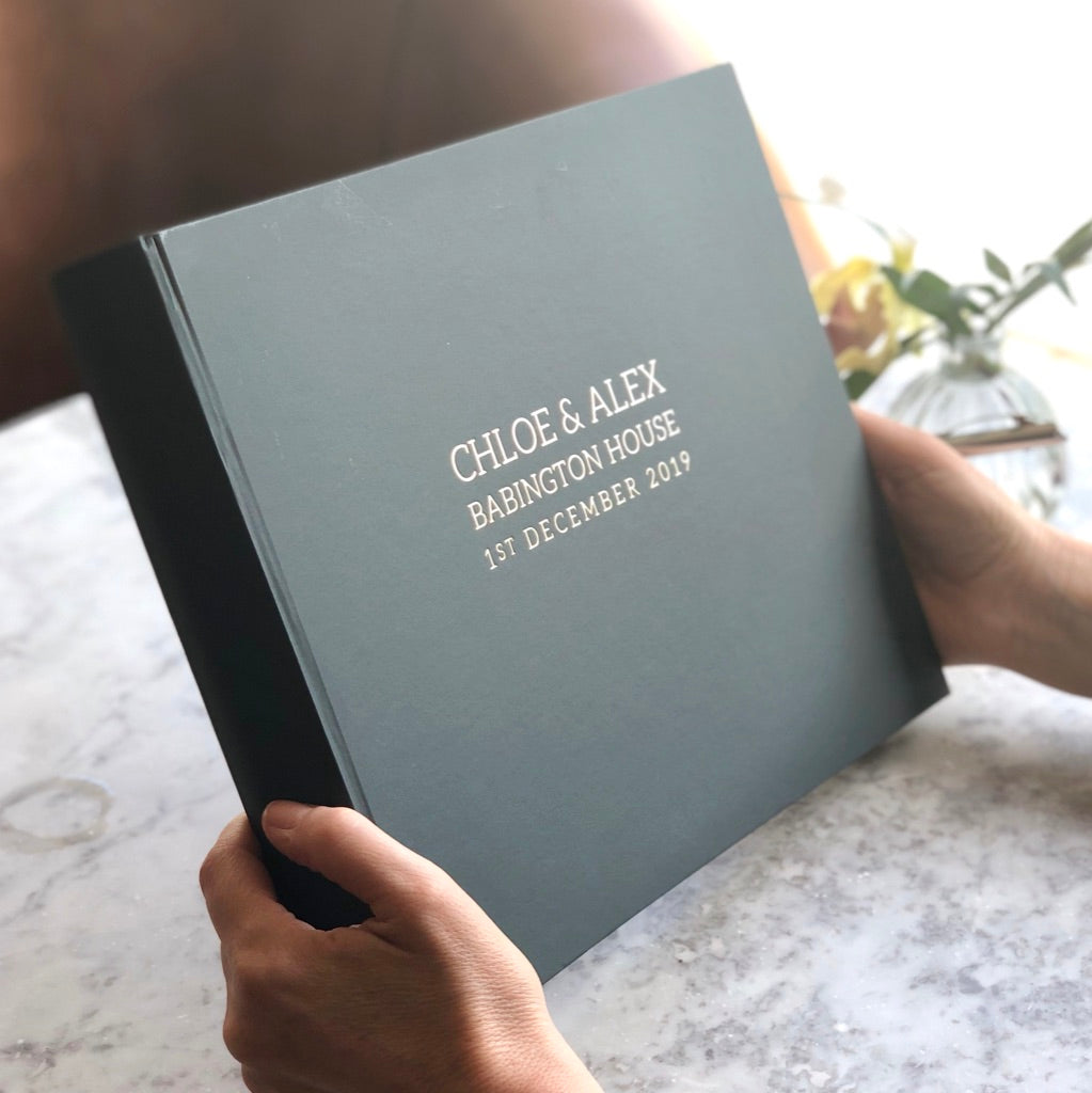 A large black guest book which has been personalised with wedding details is being held on a marble table. You can see the front and the bind of the book.