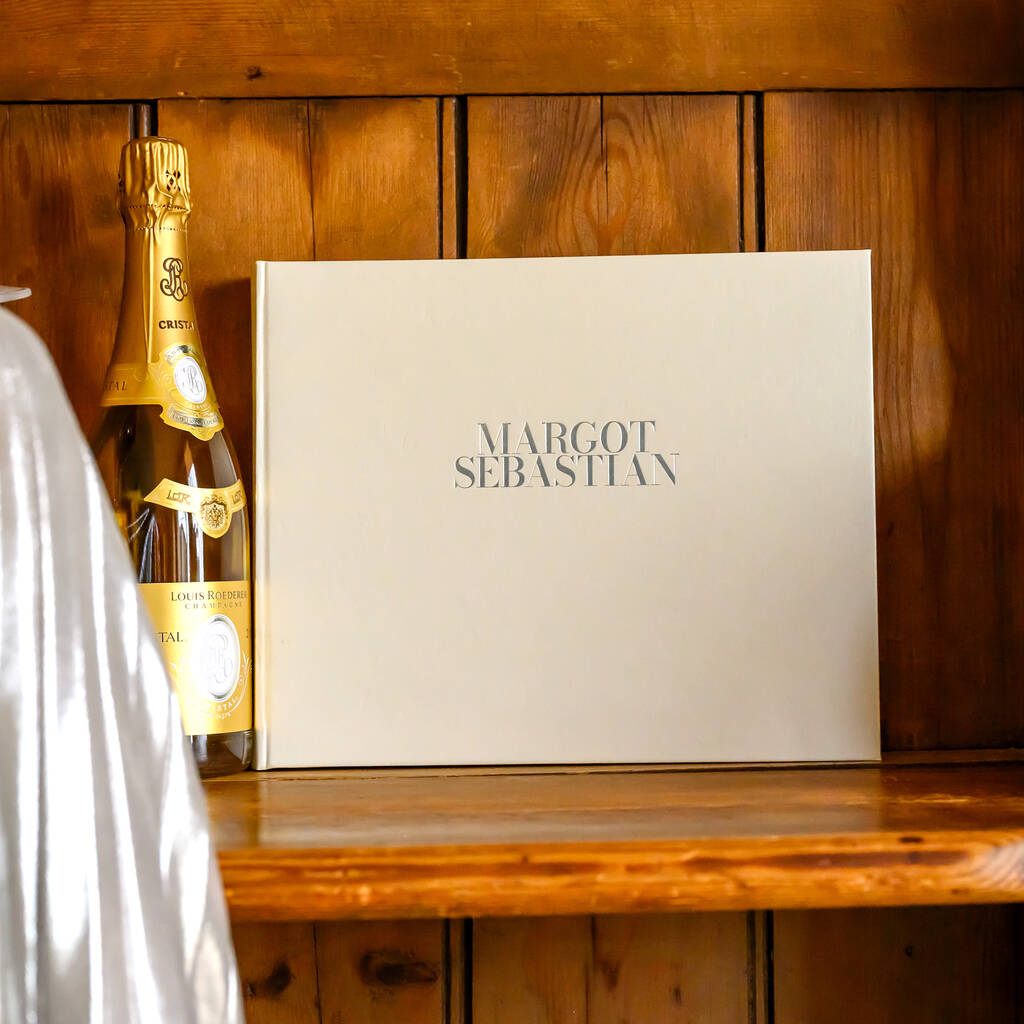 propped up against  a church pew there is a white wedding guest book. The guest book has two names written on the front in gold letters. By the side of the book there is a bottle of Crystal CHampagne.