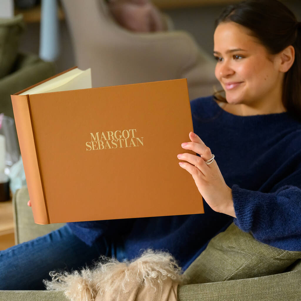 A woman in a blue jumper is sitting in an armchair. In her arms she's holding a large brown coloured wedding guest book that has been personalised on the front with two names. The names have been printed in gold foil.