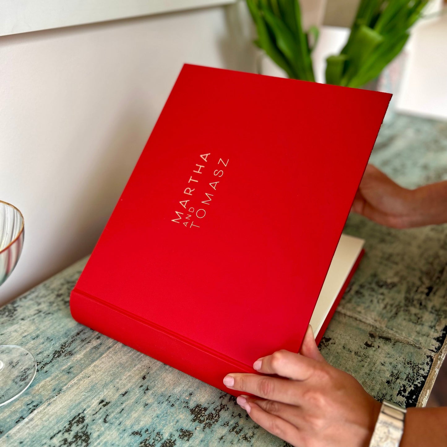 There is a woman opening a red guest book at a celebration. There is a champagne glass and a plant on the countertop.