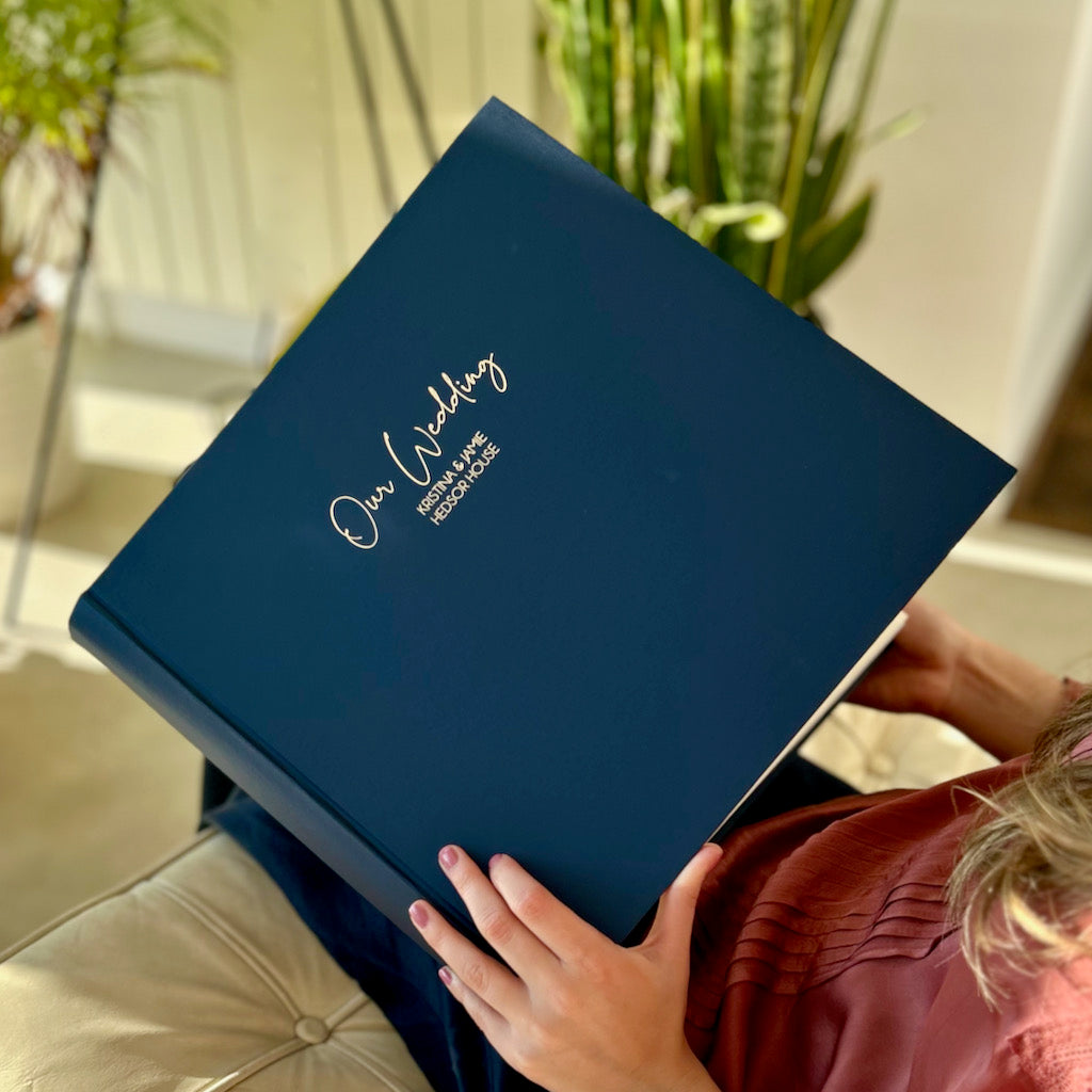 A lady is sitting on a sofa about to look through a photograph album with the title "Our Wedding" embossed on the front