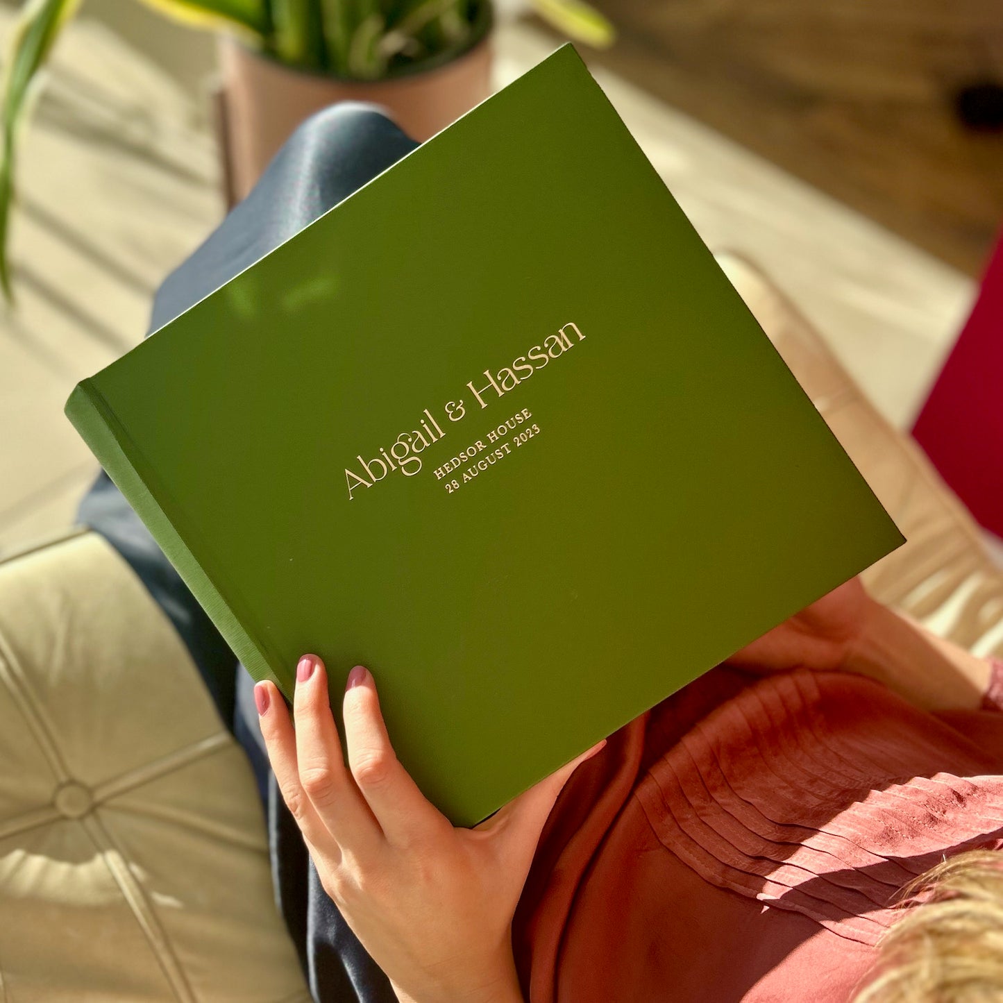a green wedding guest book is held in a young woman's hand as she sits on a sofa. The wedding guest book has golld writing on the front and details of a wedding 