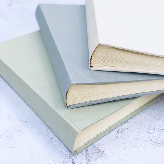 three linen wedding guest books are lying on top of each other and you can see the pages inside. There is a pale green wedding guest book, an iced grey one and an ivory one
