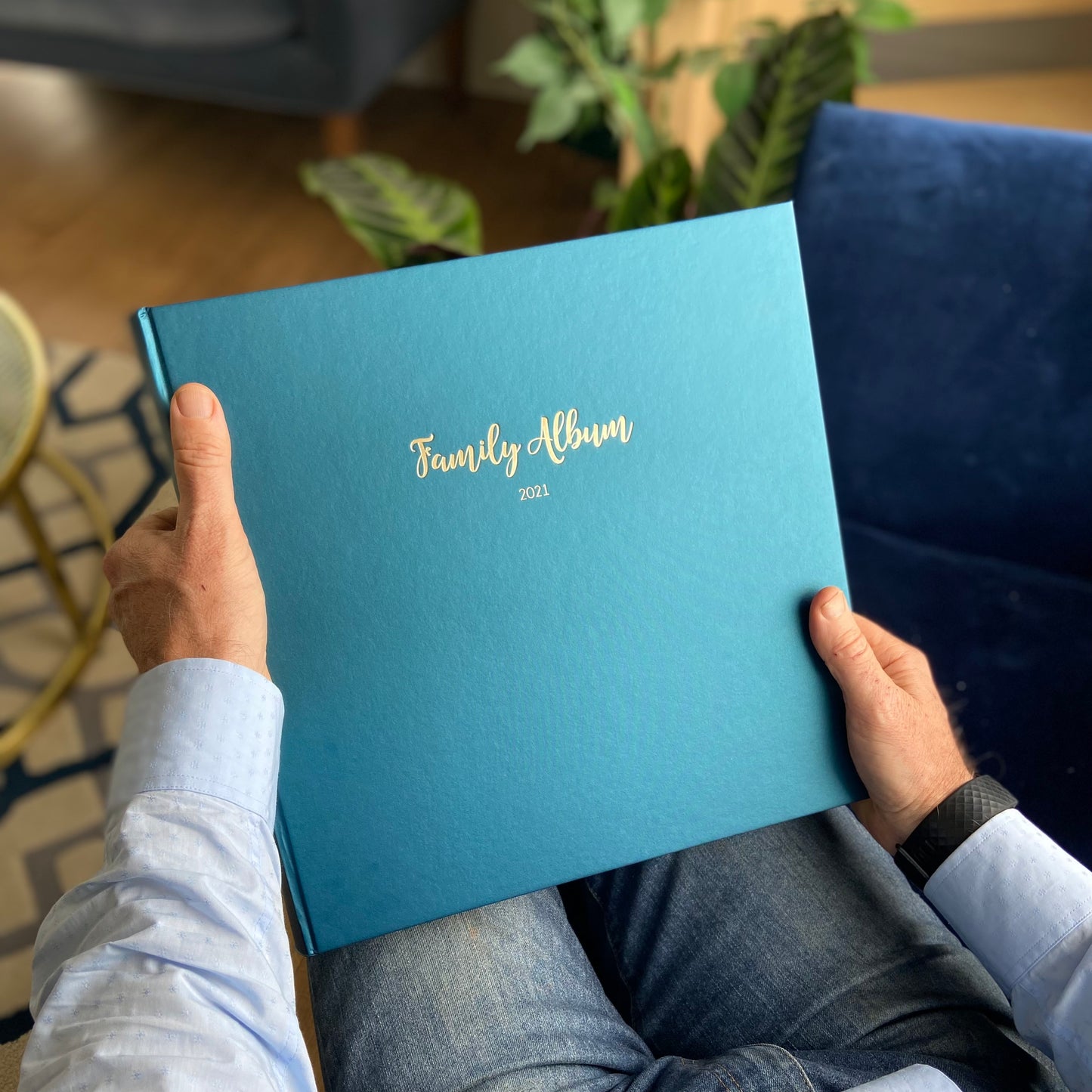 a man holds a large blue family photo album in his hands. He is sitting on a blue velvet sofa