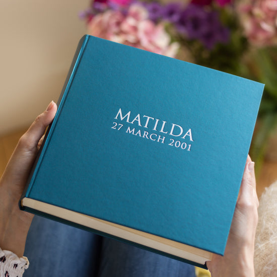 a close up image of a blue photo album which has been personalised with someone's name and date of birth. A large bunch of flowers is in the background