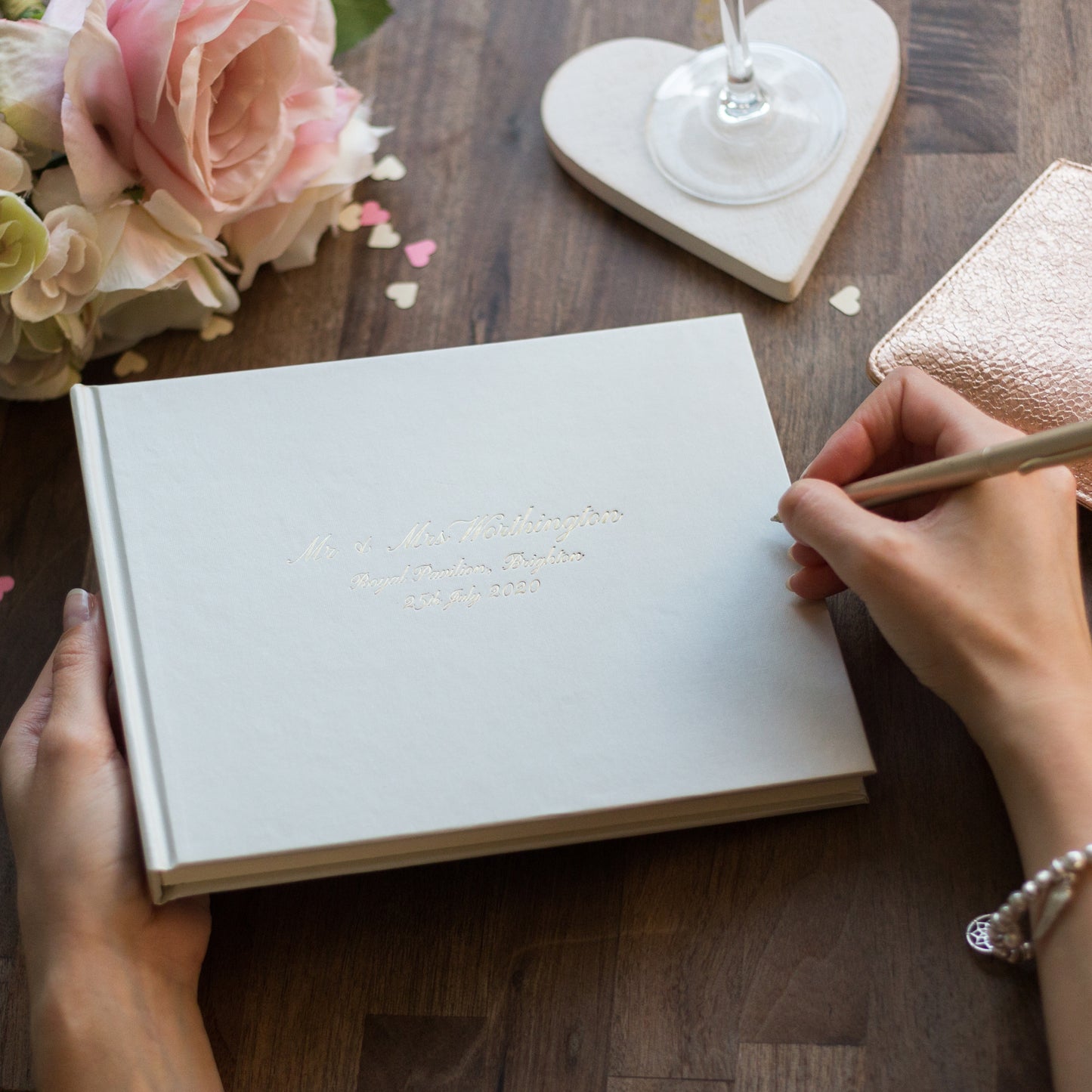 a wedding guest book is on a table and someone is about to sign it. The wedding guest book has been printed on the front with a calligraphy font. the printing says someone's wedding details 