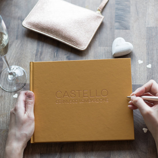 on a wooden table lies a yellow guest book which has been printed on the front with details of a house. There is also a bag on the table and a wine glass