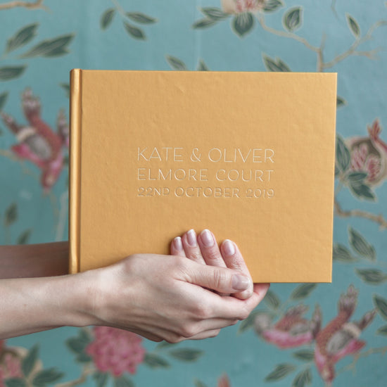 a gold wedding guest book is being held up against a green flowery back drop. The gold guest book has been pritned with someone's wedding details in a crisp font 