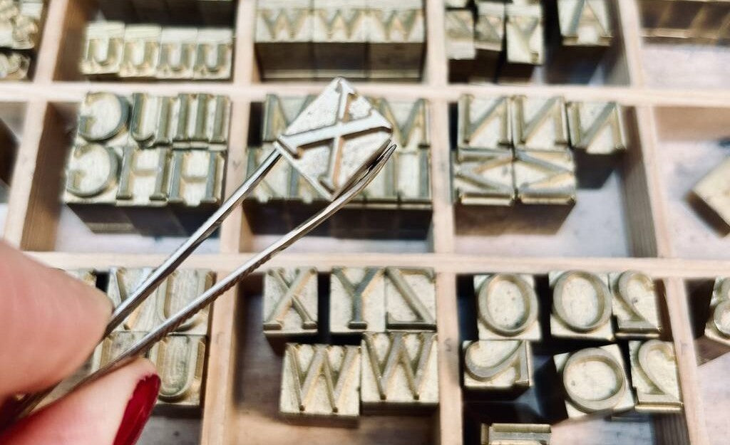 A close-up photo of brass letters used in hot foil printing and typesetting. A woman's hand is shown picking up one of the letters with a pair of tweezers.