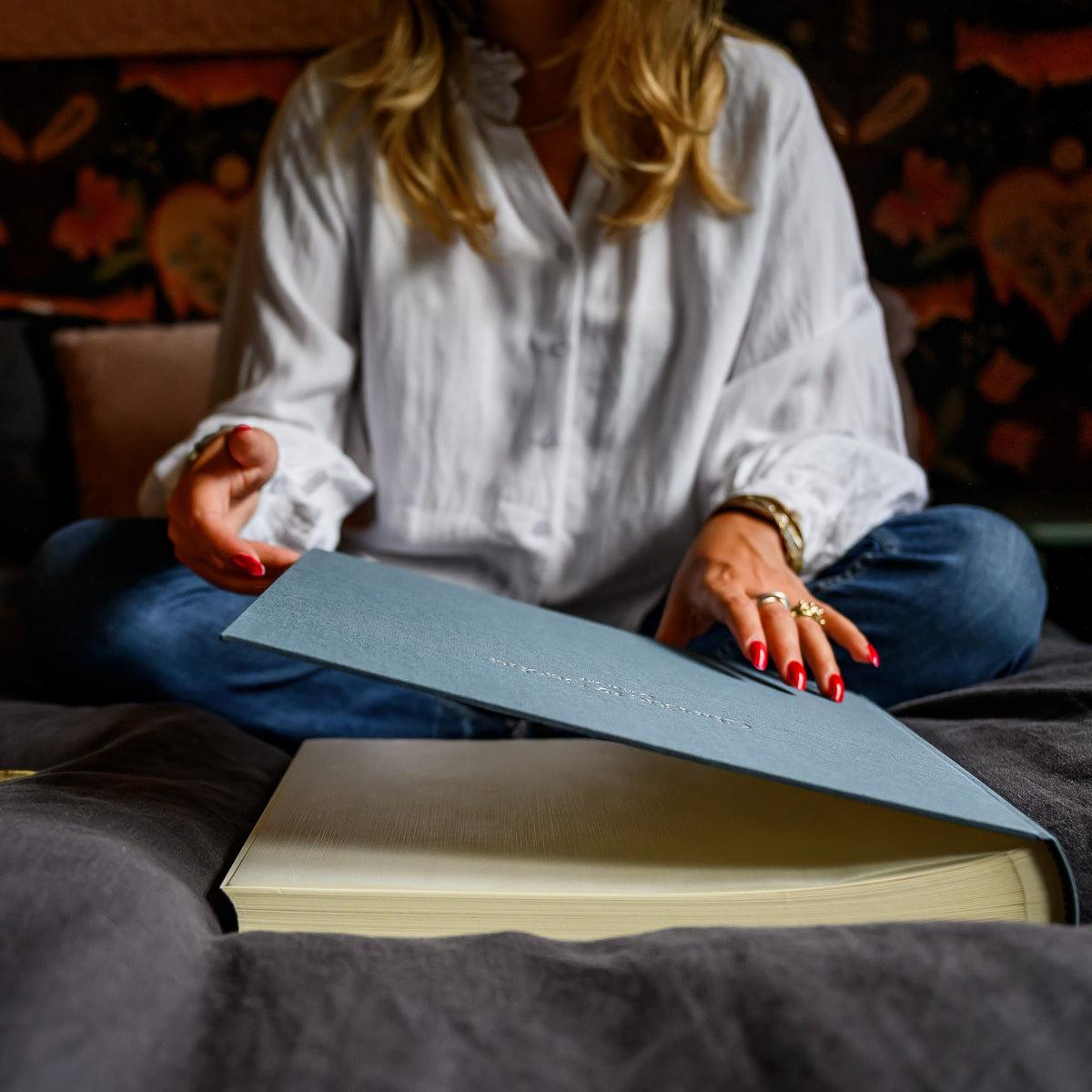 a woman is sitting on a bed looking at her grey linen wedding album.