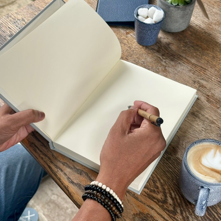 A person's hands open a blank cream-colored hardcover journal on a rustic wooden table, holding a wooden pen. A cup of latte and other desk accessories are visible in the background.
