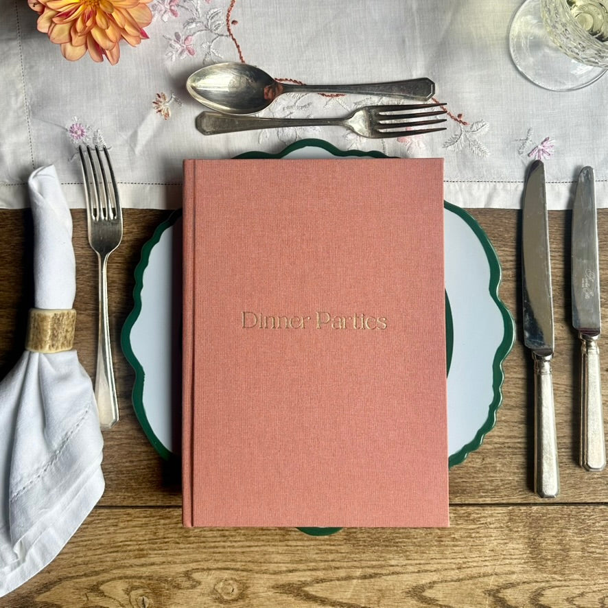 A hardcover book titled 'Dinner Parties' in gold lettering, standing upright on a green-rimmed pale blue plate in a styled dining flat lay with silver cutlery, a folded white napkin, and a floral tablecloth.
