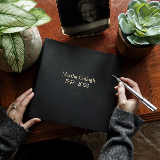 on a wooden table lies a black condolence book which has been printed with a woman's details in gold letters. There are some plants on the table and a photo of the deceased