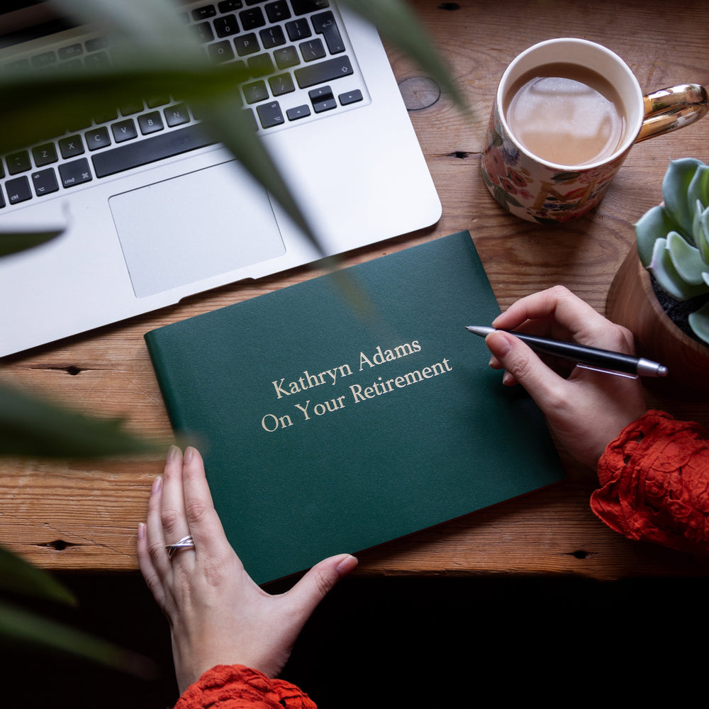 a green retirement book is on a work desk and someone is about to write in it. The retirement book has been printed with details of a work colleague. 