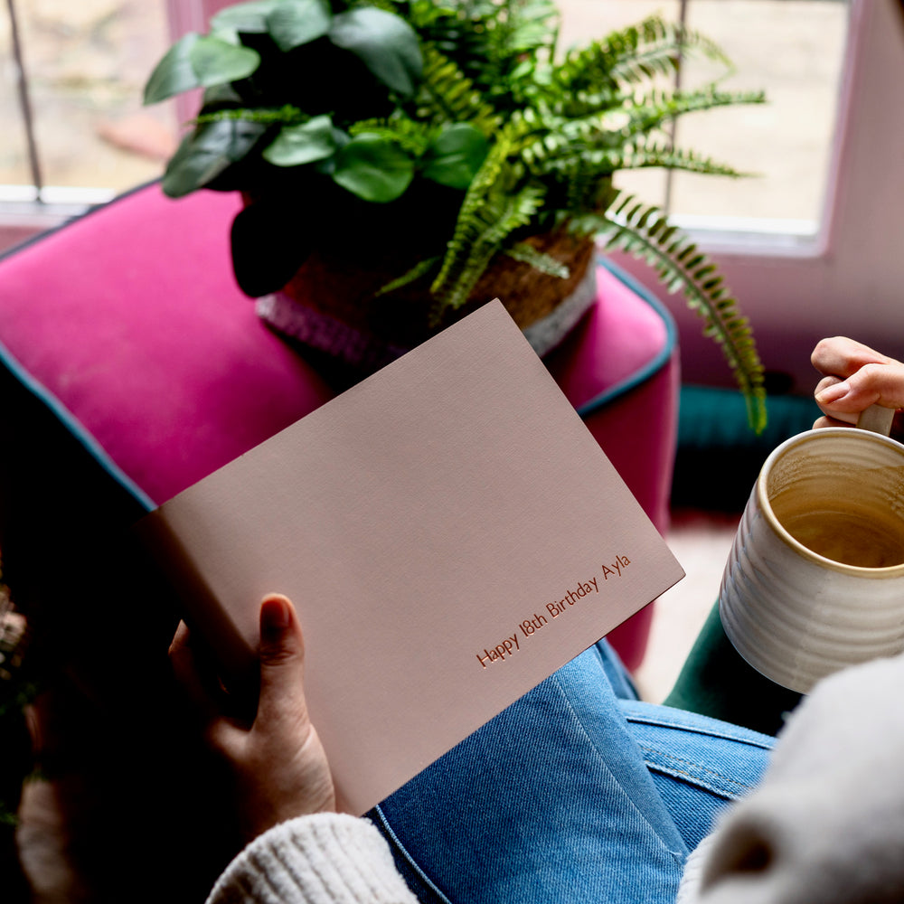 a woman wearing a pair of jeans is sitting in a chair holding a small photo album in one hand a cup of coffee in the other