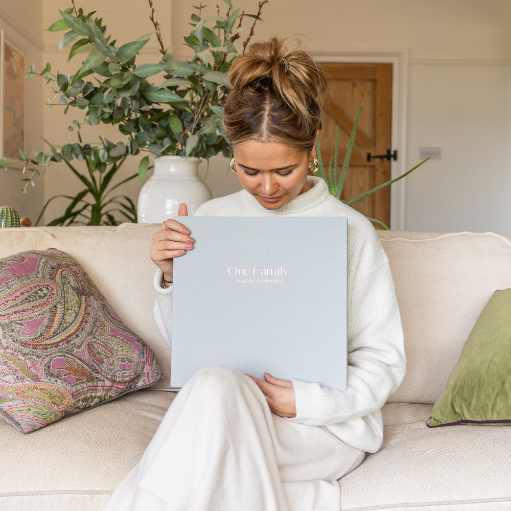 
                      
                        Woman sitting on  a sofa holding a grey linen photo album, printed with 'Our Family: A Lifetime of Memories'
                      
                    