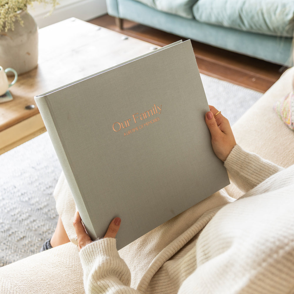 Person holding a grey linen photo album personalised with the words 'Our Family' in a cozy living room.