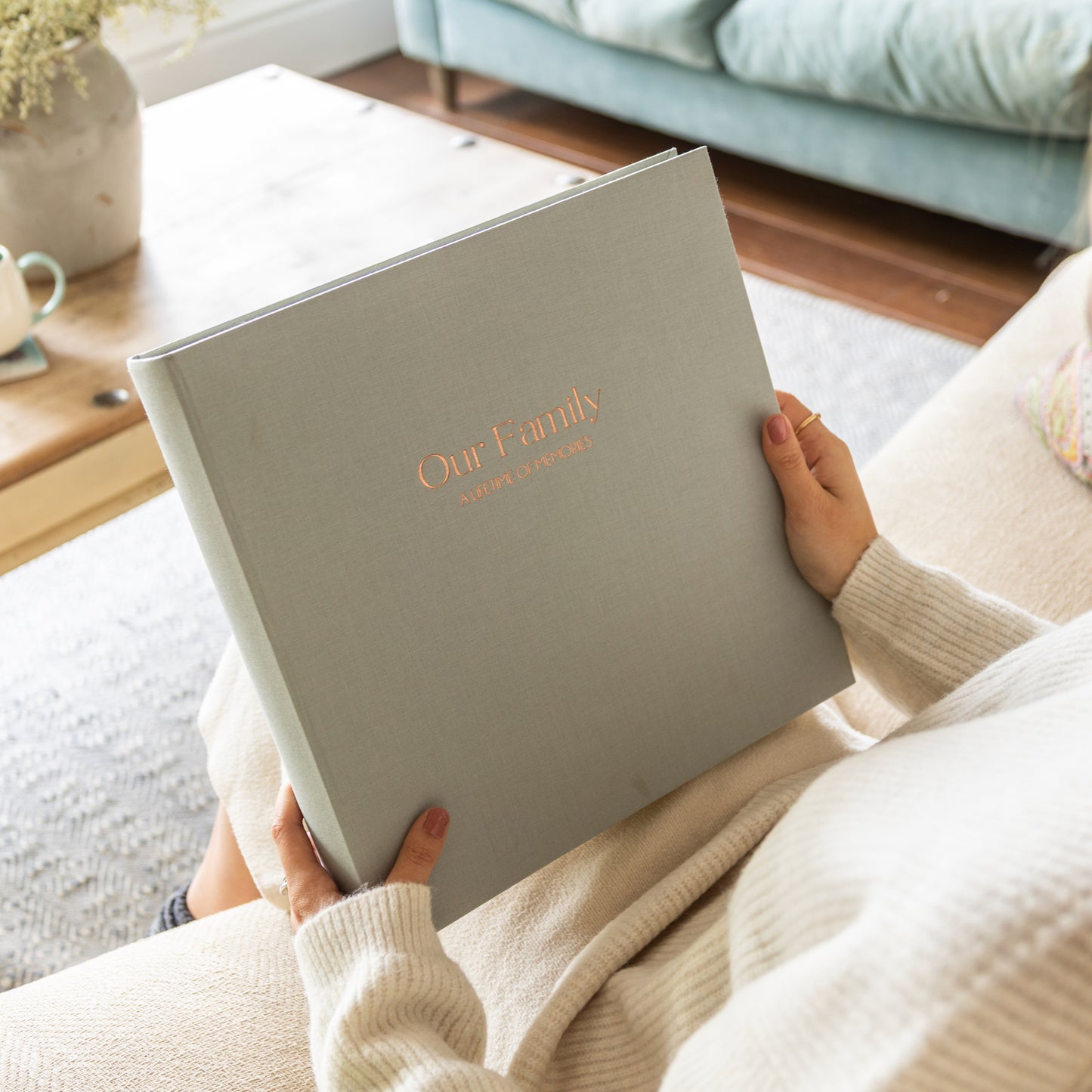 Person holding a grey linen photo album personalised with the words 'Our Family' in a cozy living room.