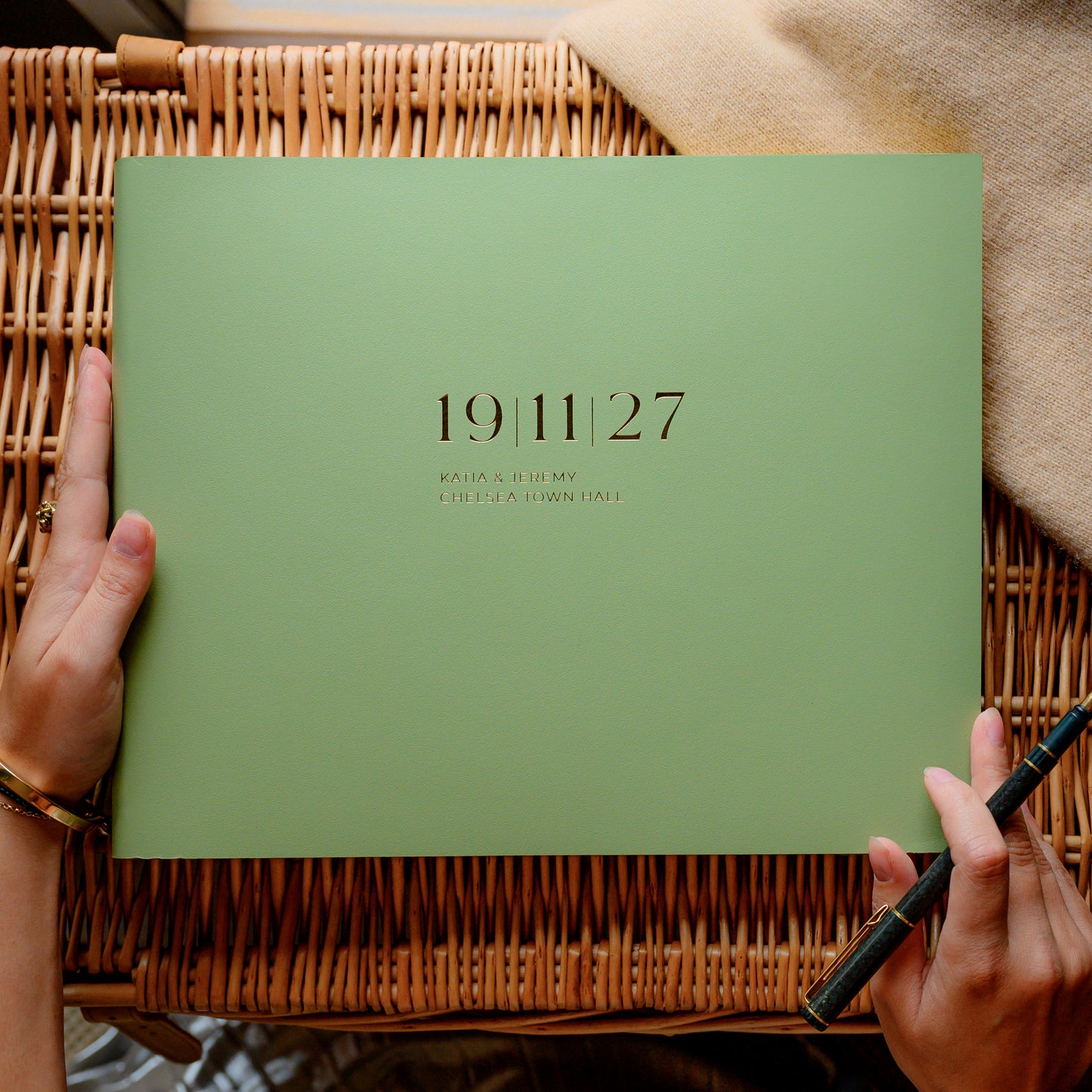 Sage green wedding guest book with wedding date and details printed on the cover, held by a person over a wicker surface.