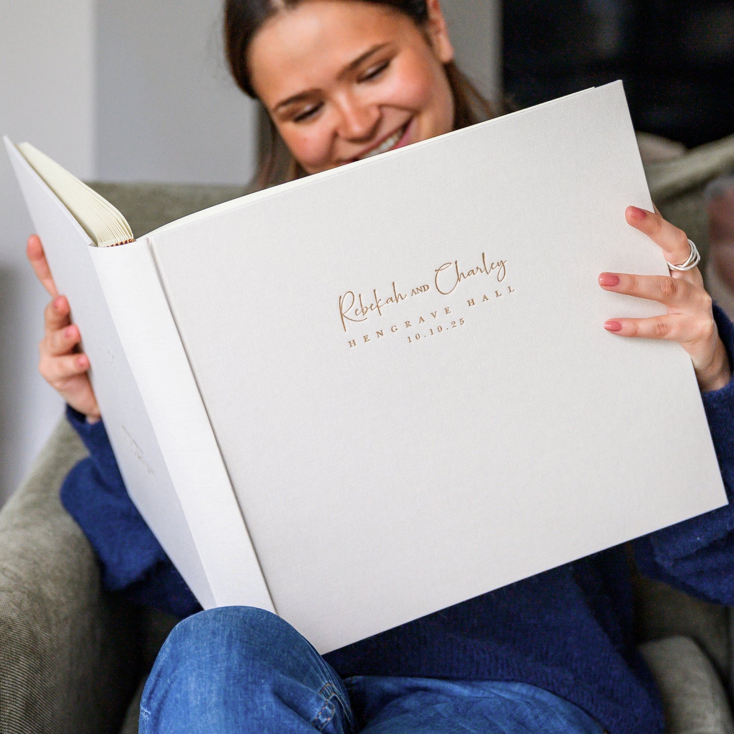 a woman is sitting in a chair holding a really large ivory linen wedding album. The woman is dressed all in blue. The wedding album has been printed with gold lettering on the front.