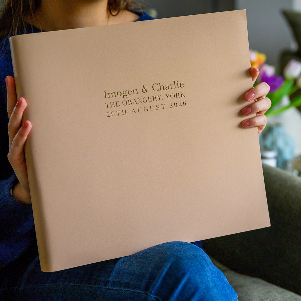 A woman is sitting in a chair holding a large wedding album in her hands. The album has been personalised with wording, In the background there are some flowers in a vase and a cup of coffee.