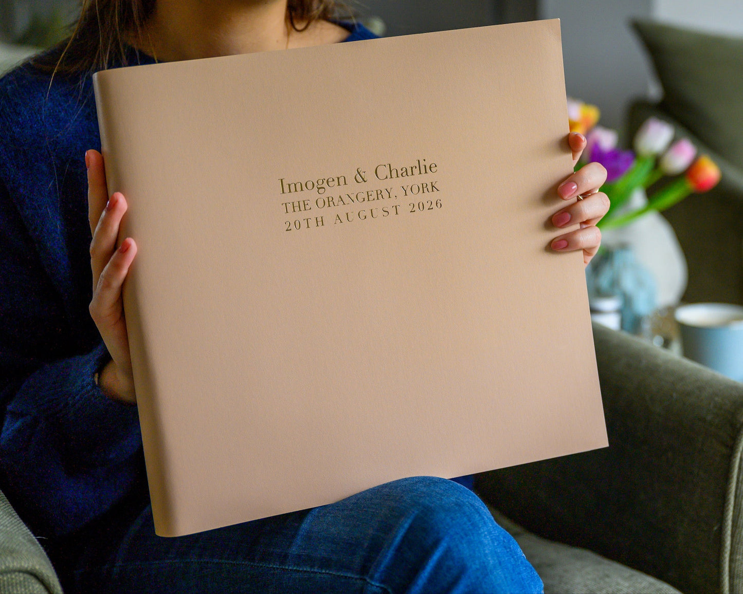 A woman is sitting in a chair holding a large wedding album in her hands. The album has been personalised with wording, In the background there are some flowers in a vase and a cup of coffee.