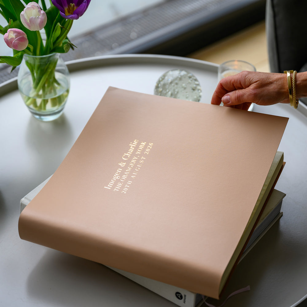 on a white coffee table there is a large pink photo album being opened by  a hand. In the background there is a glass vase and a paperweight. The wedding photo album has been personalised with gold wording 