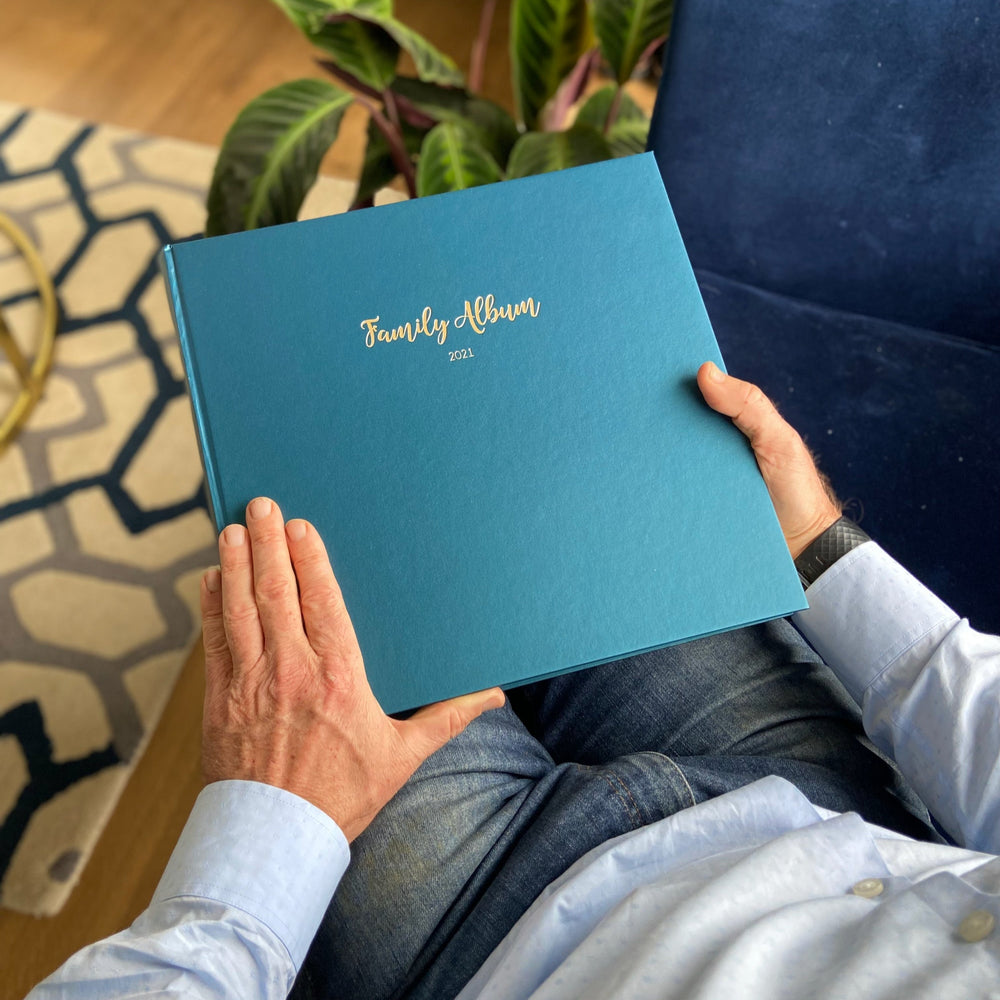Person holding a peacock green family photo album personalised with gold text in a home setting.