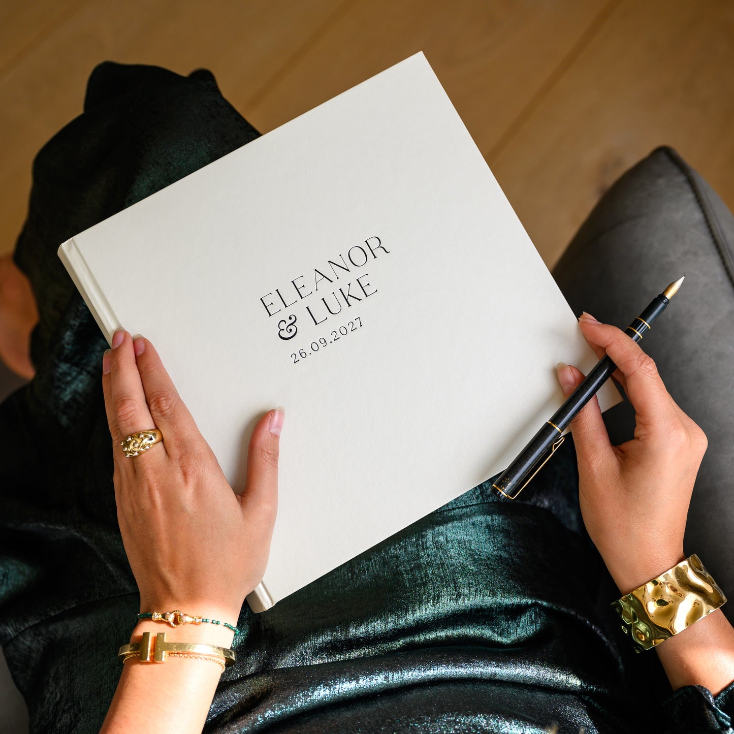 Woman holding a hardback wedding guest book printed with 'Eleanor & Luke' and a pen, wearing gold jewelry.