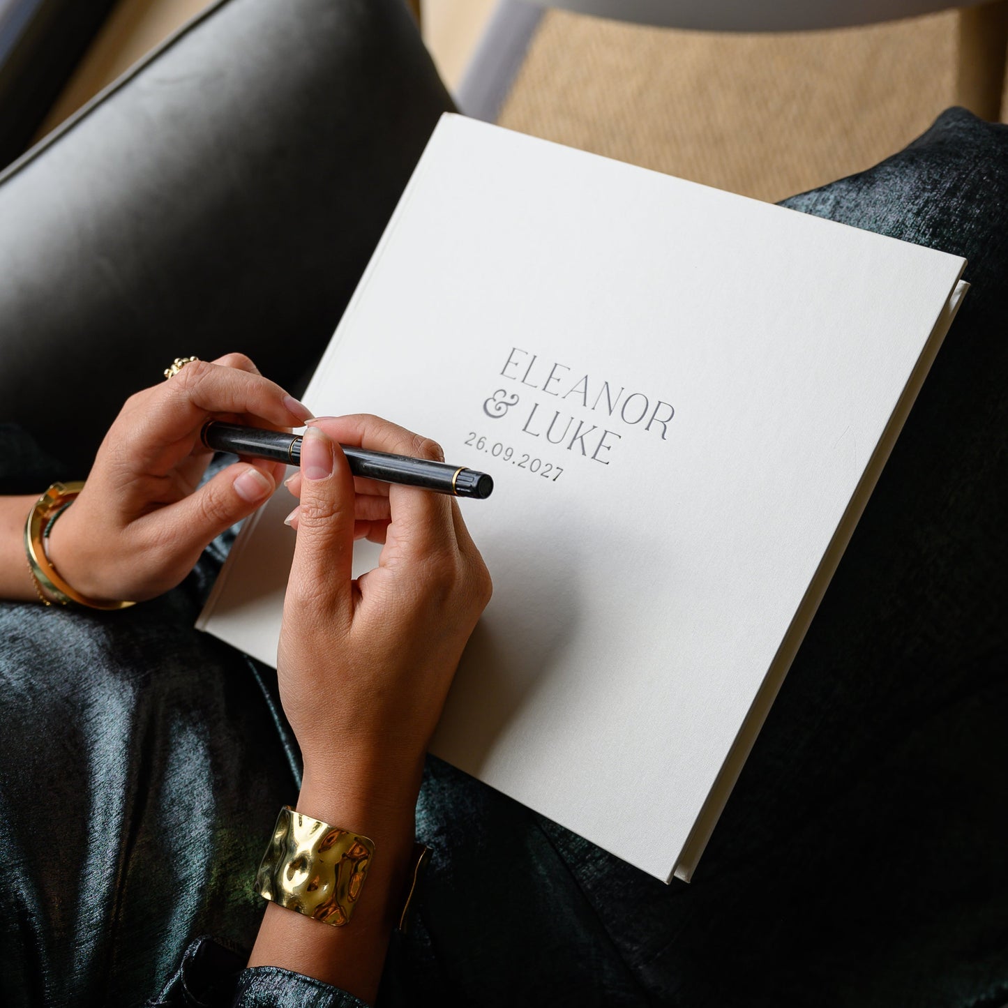 Woman holding a pen over a white wedding guest book printed with 'Eleanor & Luke' and a date.