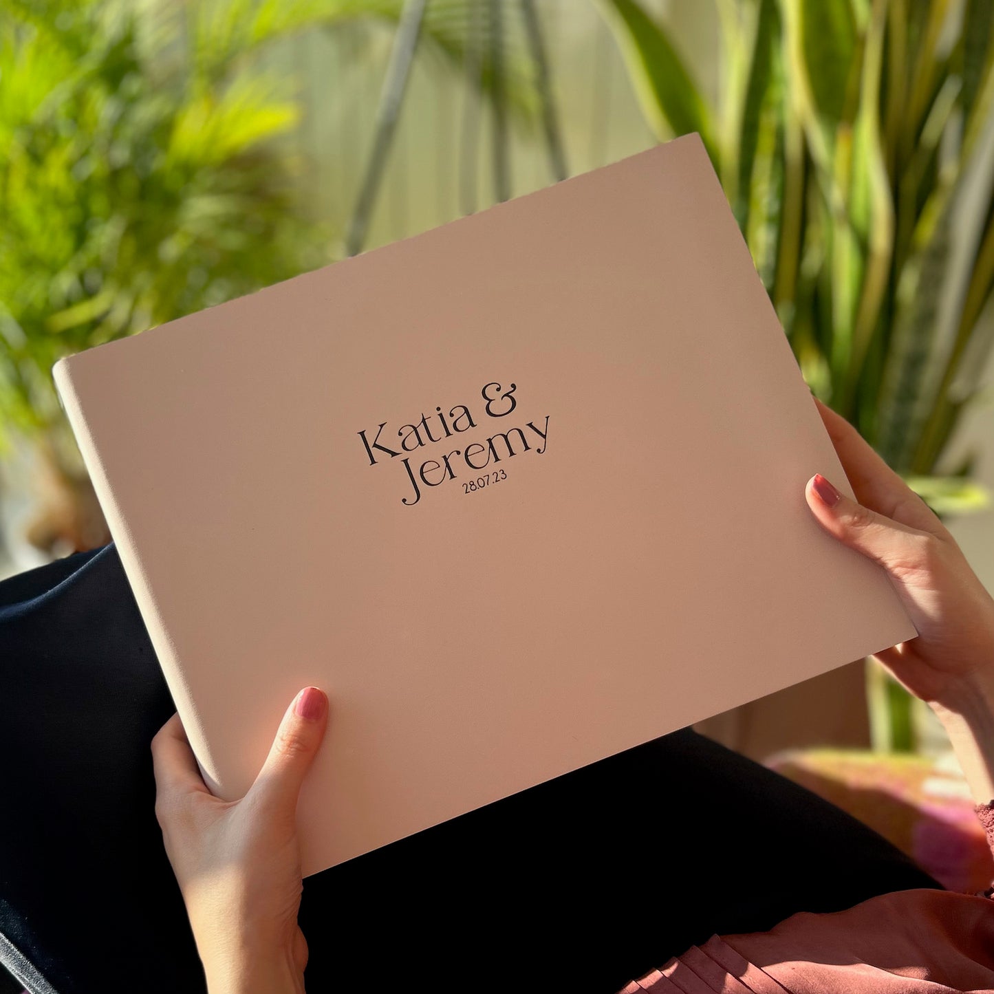 a blush pink landscape wedding guest book is being held by a woman in a chair. On the front of the wedding guest book is the names of the bride and groom in black letters. There are plants in the background