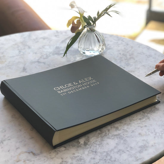 a large wedding guest book is laying on the table. It has gold lettering on the front. Someone is holding a pen as if they are going to write in it