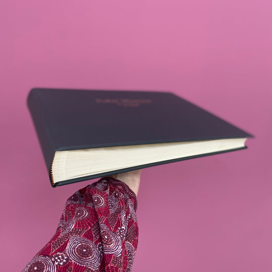 a black wedding guest book is being held up against a pink wall so you can see the traditional binding, The landscape guest book has 40 pages of photoboard