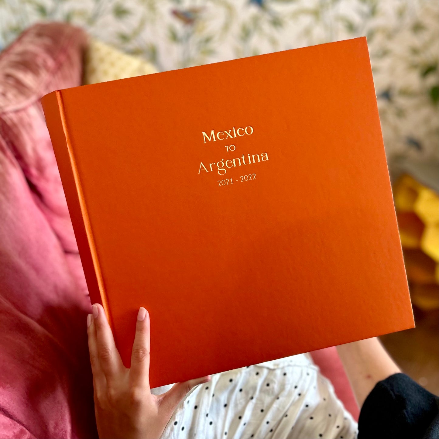 a woman is sitting on a sofa holding a large orange photo album which has been printed on the front with a holiday destination