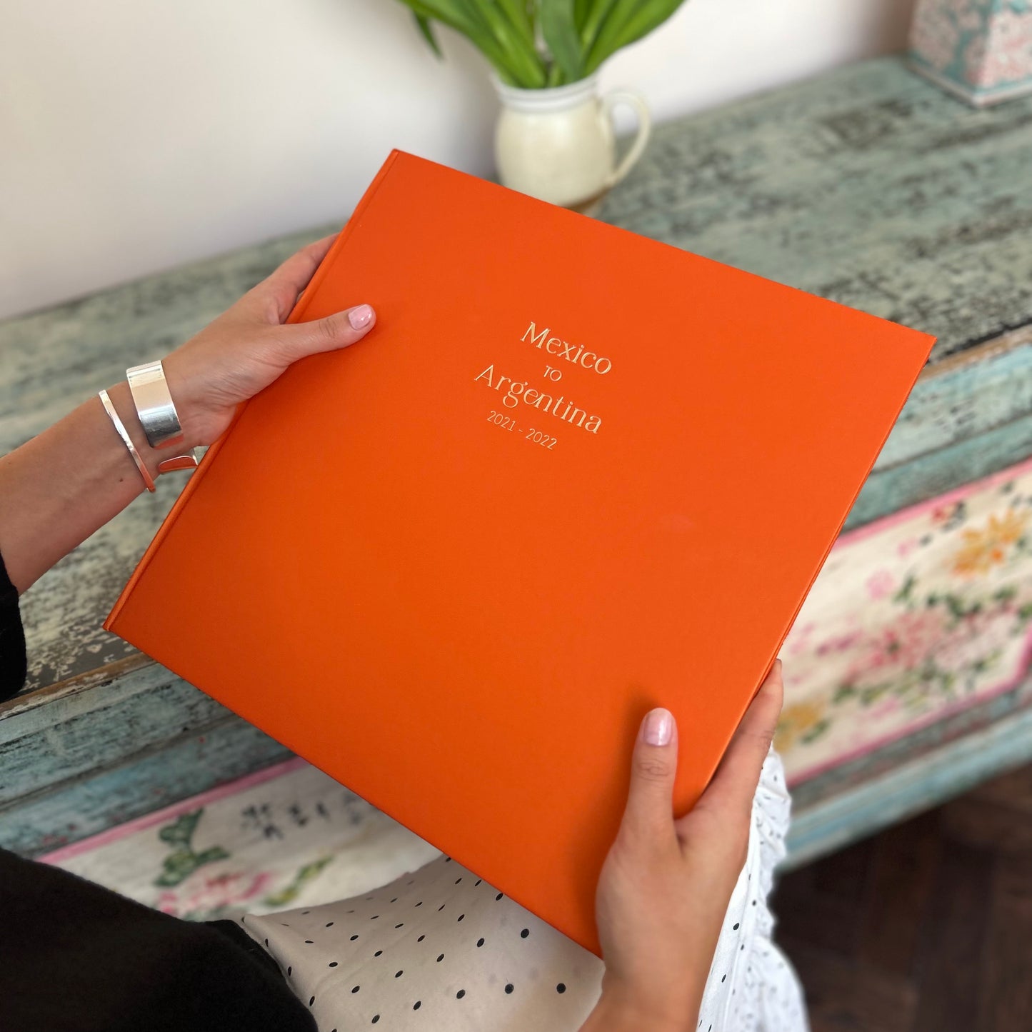 a woman holds a bright orange photo album with a holiday destination written on the front. The writing is in gold foil