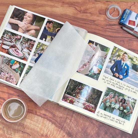 a large wedding photo album lies open on a wooden table and inside it is full of wedding photos. There are also some mounting squares also on the table 