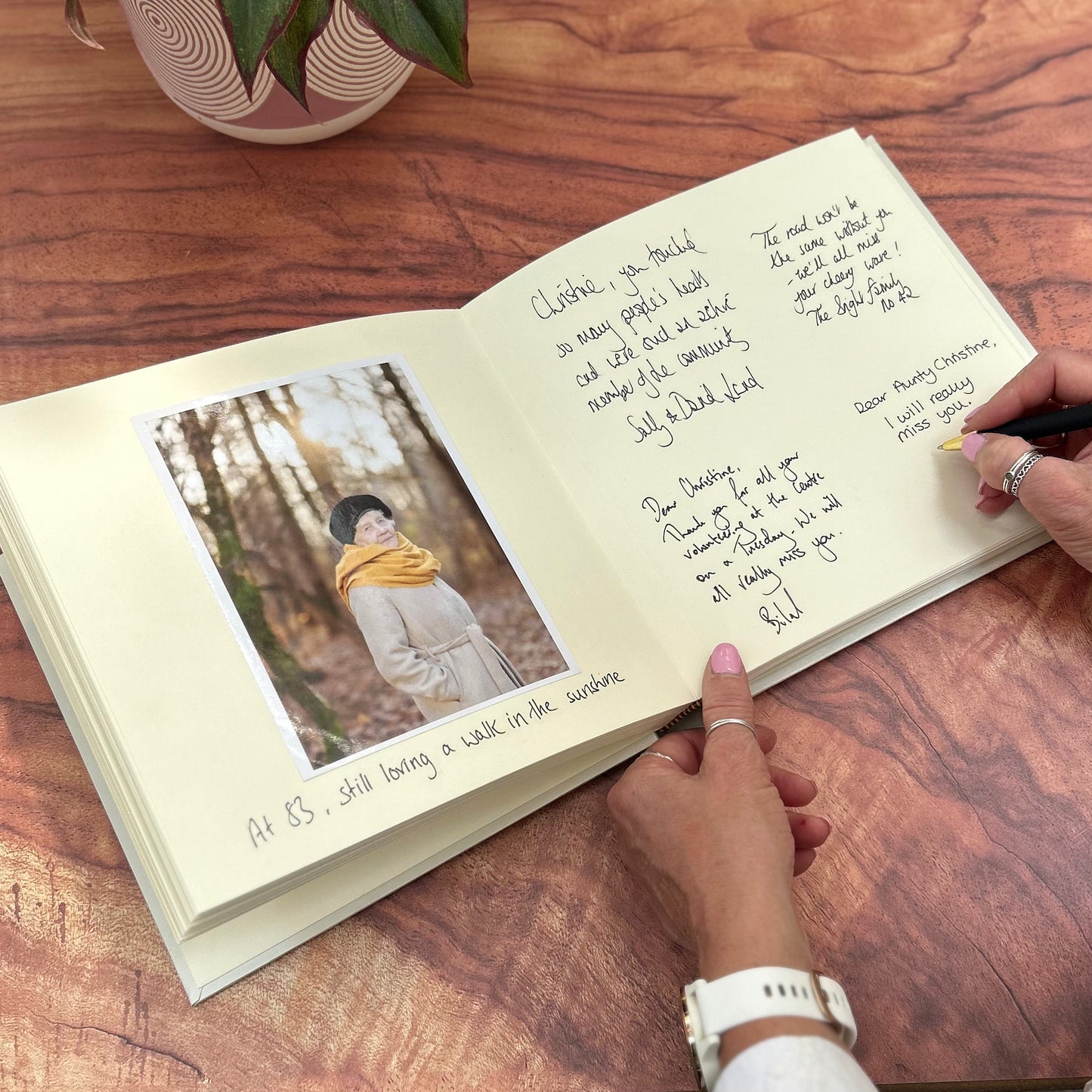 There is a woman writing a message in a condolence book. There are lots of messages already inside and a photograph of the loved one who has passed away. 