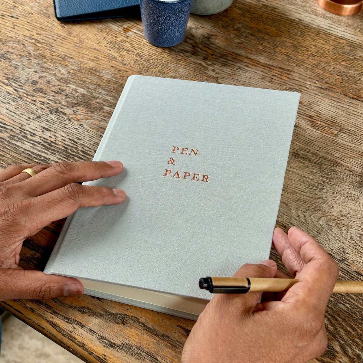 A light grey linen hardcover journal with 'PEN & PAPER' printed in copper-toned text on the cover, held by two hands over a rustic wooden table, with a pen in one hand.