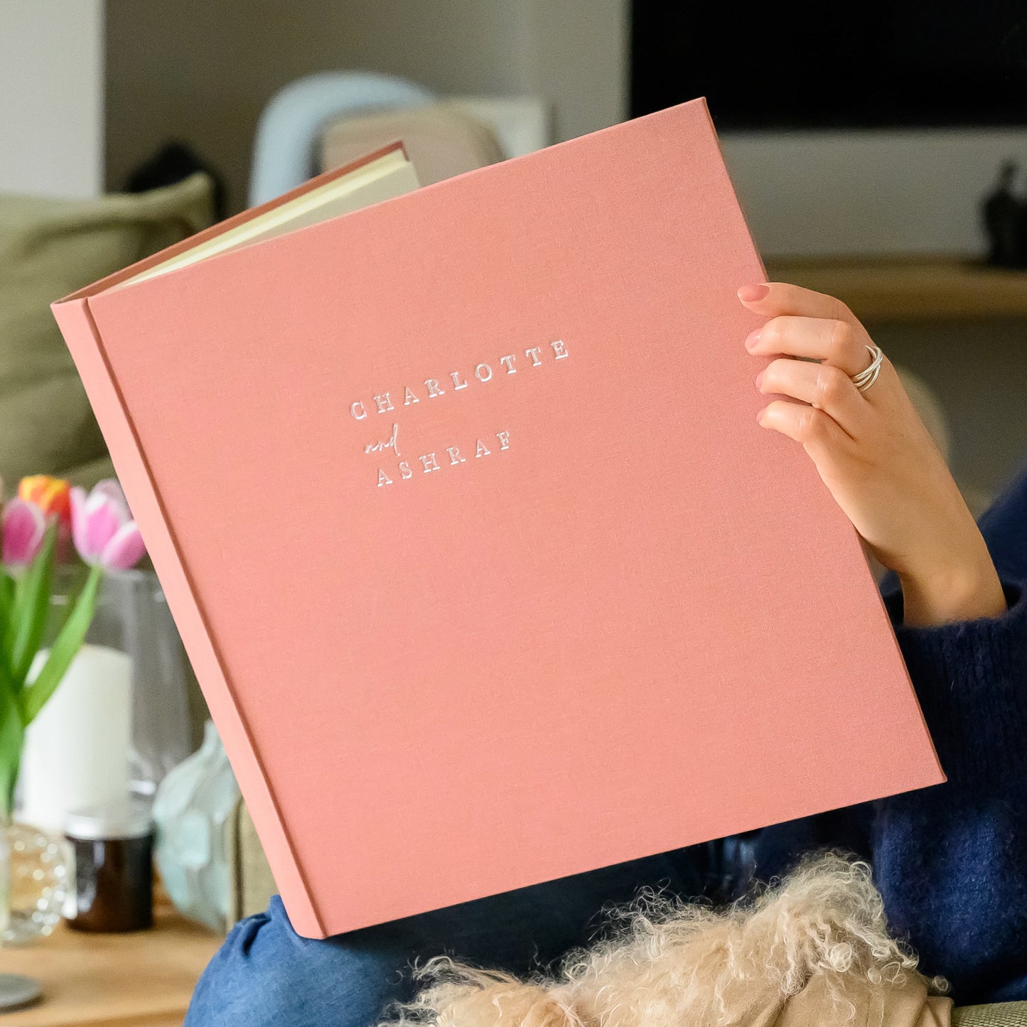 Person holding an antique rose wedding album with 'Charlotte and Ashraf' printed on it, in a home setting.