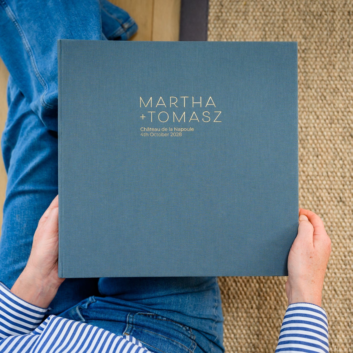 A woman sits holding a large, grey linen wedding photo album, printed in gold with two names and wedding details
