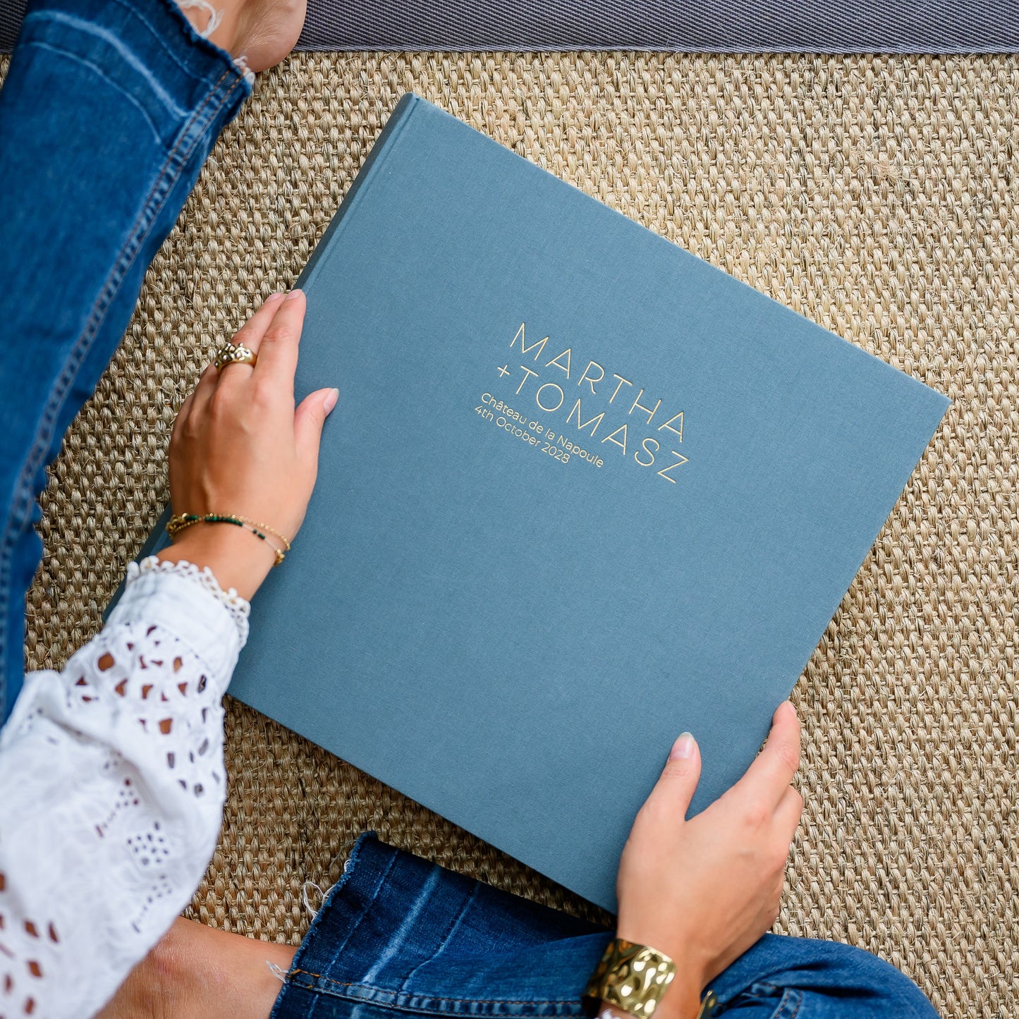 A woman holds a large, grey linen wedding photo album, printed in gold with two names and wedding details
