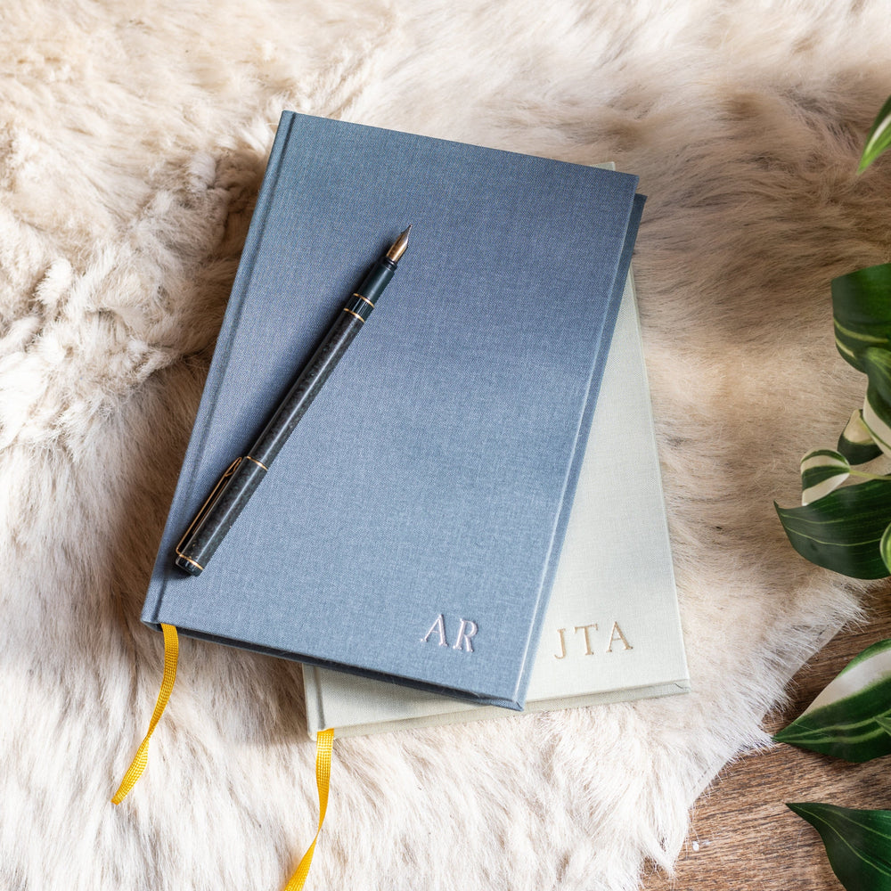 A pile of linen-bound diaries, personalised with printed initials, with a pen resting on top.