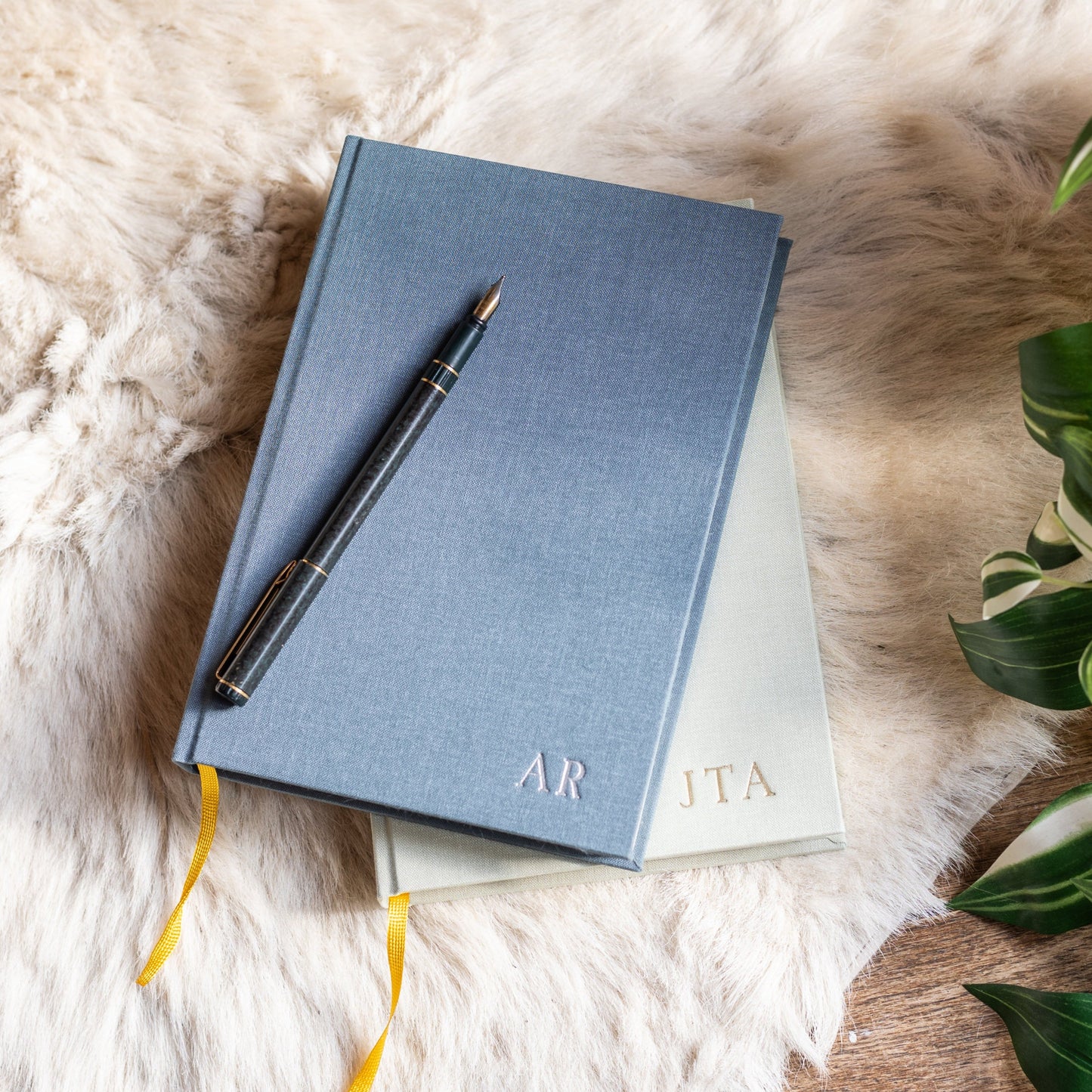 A pile of linen-bound diaries, personalised with printed initials, with a pen resting on top.