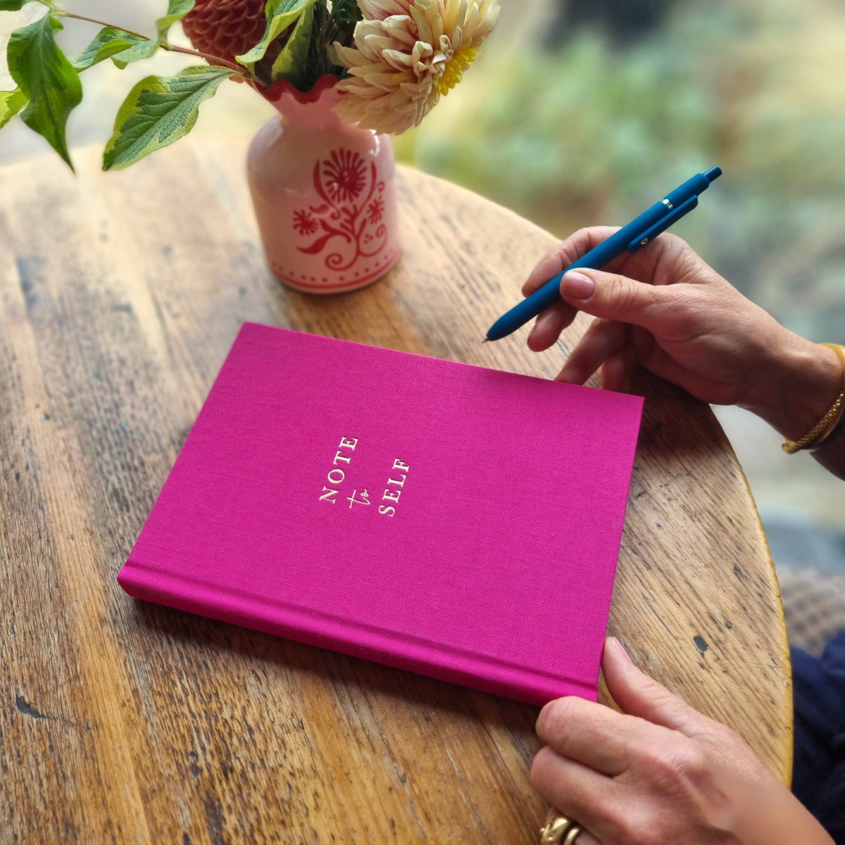 A bright pink hardcover journal with gold foil print reading 'NOTE to SELF' sits on a rustic wooden table. A woman's hands are visible holding a blue pen, with a small pink floral vase in the background.