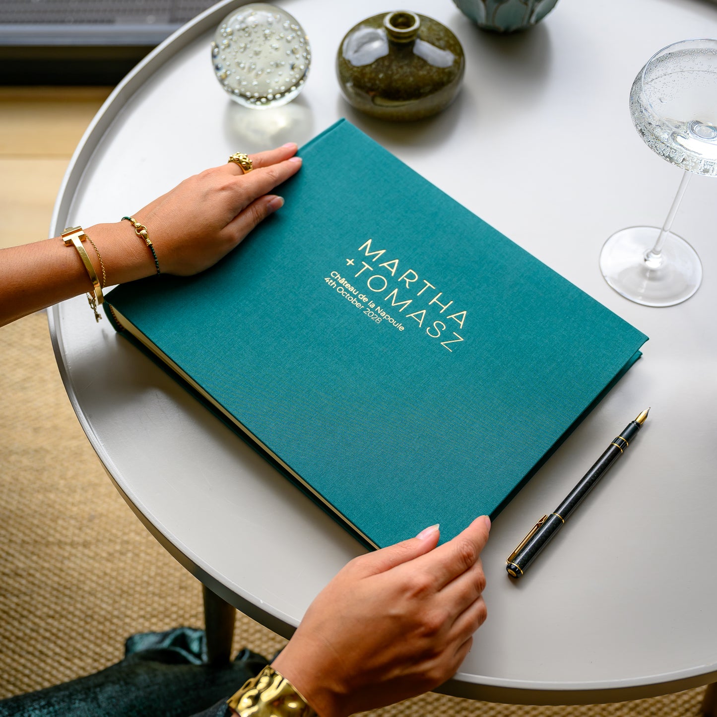 A woman opens a large, peacock green, landscape linen wedding guest book, printed in gold with two names, on a coffee table