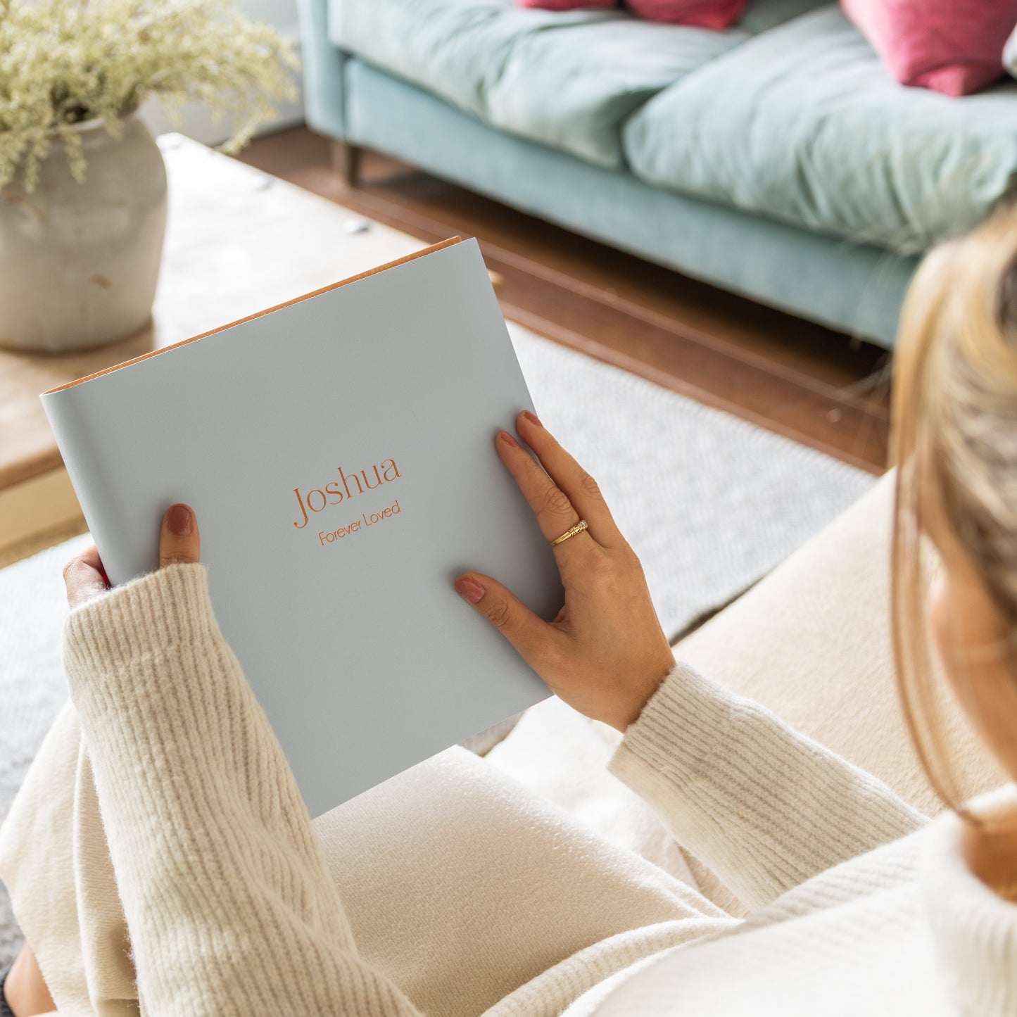 Woman holding a condolence book titled 'Joshua Forever Loved' in a cozy living room setting.