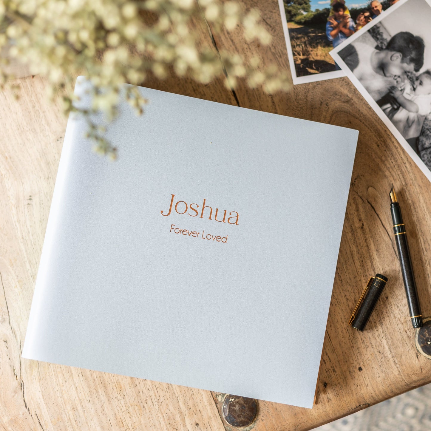 Leather Condolence book personalised with 'Joshua Forever Loved', on a wooden surface with photos and pens.