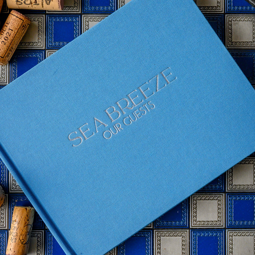 Sky blue linen-covered guest book embossed with 'Sea Breeze Our Guests', placed on a decorative surface with wine corks.
