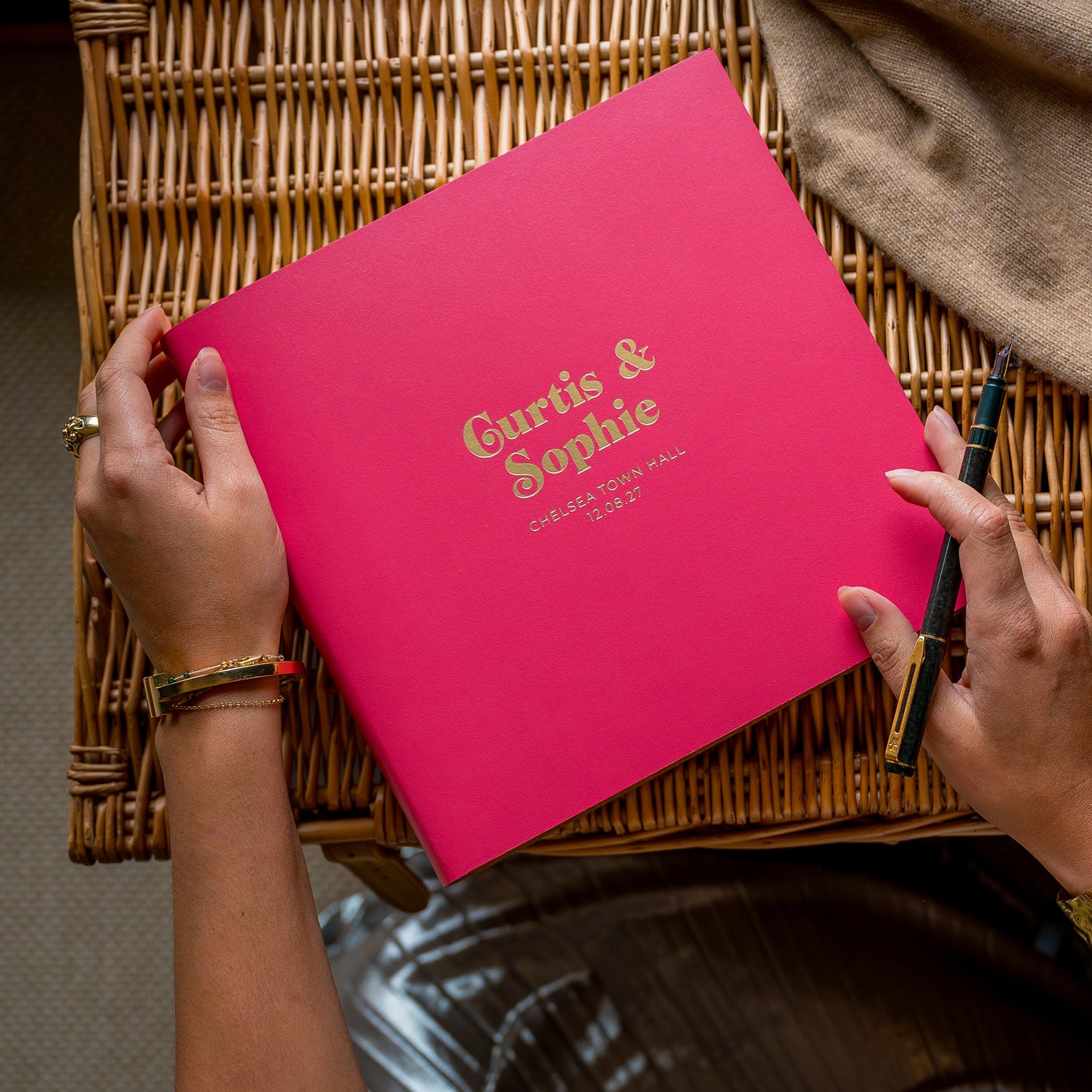 A woman holding a pink wedding guest book printed with 'Curtis & Sophie' on it, sitting on a woven chair.